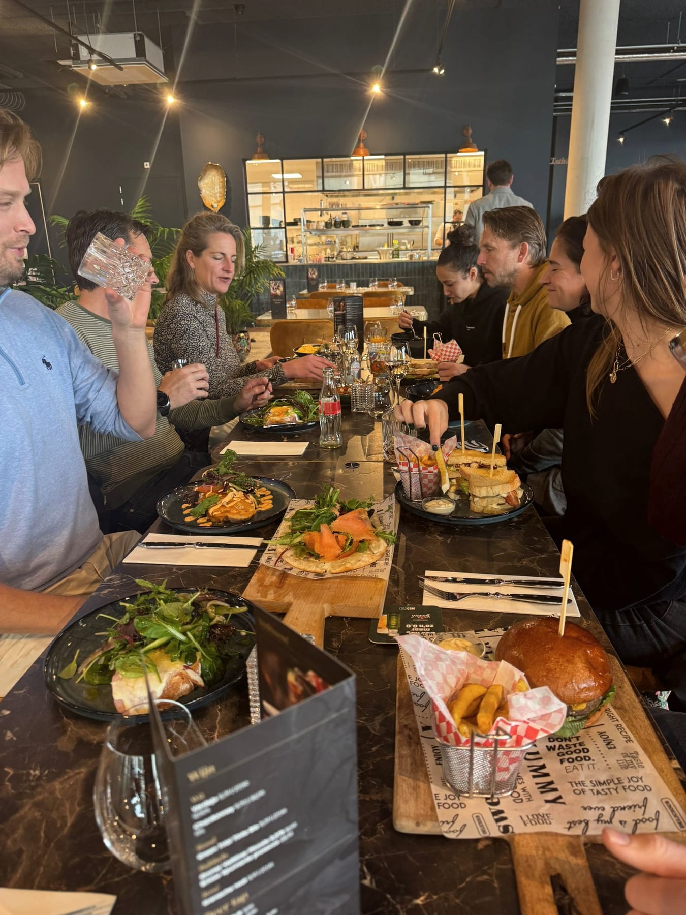 Gezelschap aan een lange tafel in Brasserie The Scene met gedeelde gerechten op planken